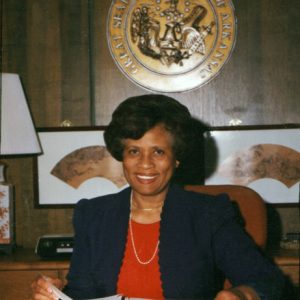 Black woman in jacket blouse at desk holding binder seal of Arkansas plaque on wall