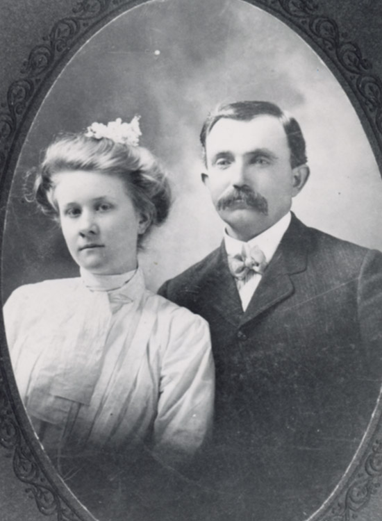 White woman with flowers in her hair and white dress next to white man with mustache in suit