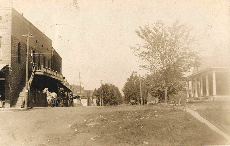 Horse drawn wagon on town street under balcony of multistory building with houses across the street and in the background