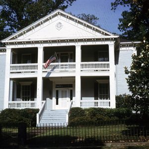 Symmetrical two-story house with covered porch and covered balcony above it with an American flag displayed from it and steps leading to the front door