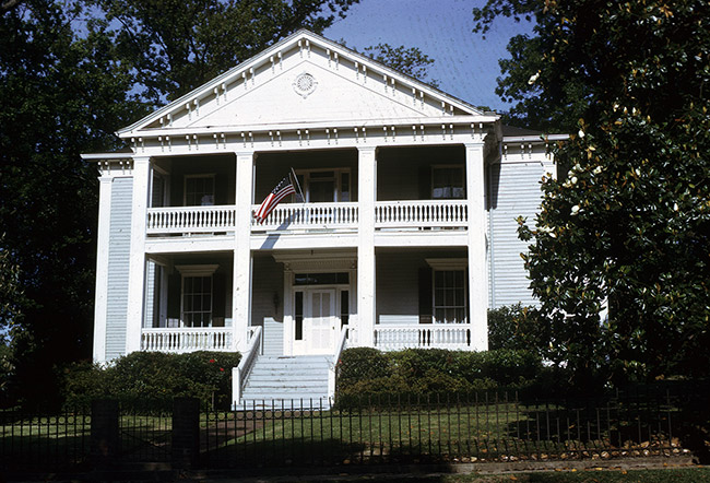 Symmetrical two-story house with covered porch and covered balcony above it with an American flag displayed from it and steps leading to the front door