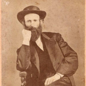 White man with long beard in hat and suit sitting in chair with fist raised to his face