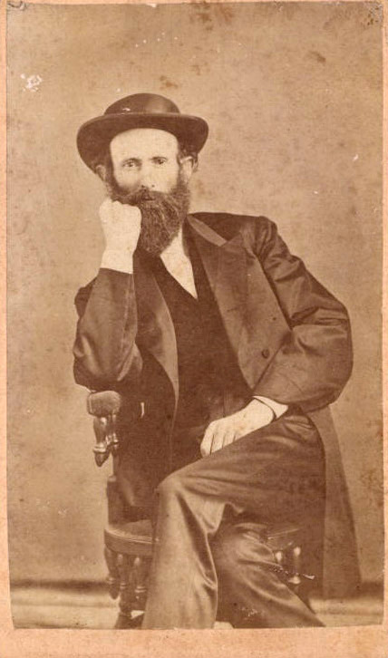 White man with long beard in hat and suit sitting in chair with fist raised to his face