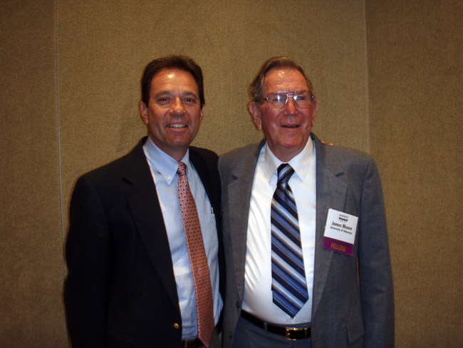 White man in black suit jacket and tie standing next to older white man in glasses and gray suit and tie
