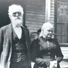 Older white man in suit and older woman in dress standing outside house with covered porch