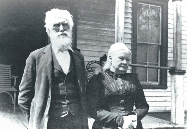 Older white man in suit and older woman in dress standing outside house with covered porch