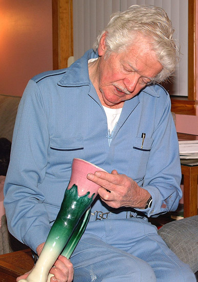 Older white man with mustache in blue shirt and pants looking at a painted glass vase