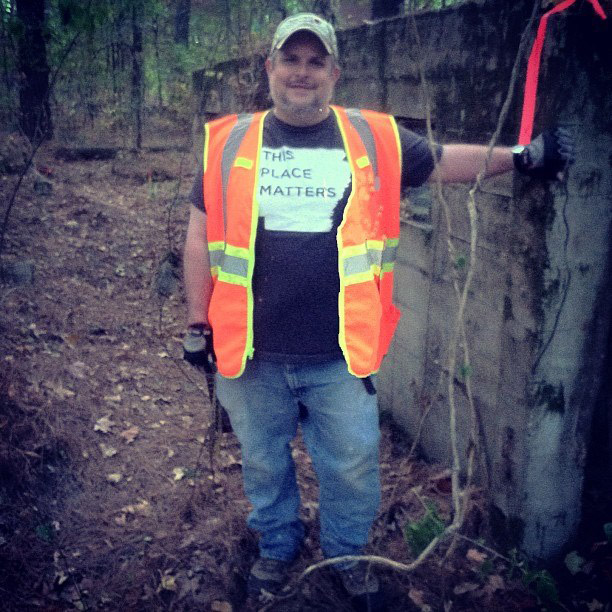 White man in cap orange vest and jeans leaning on concrete wall in forested area