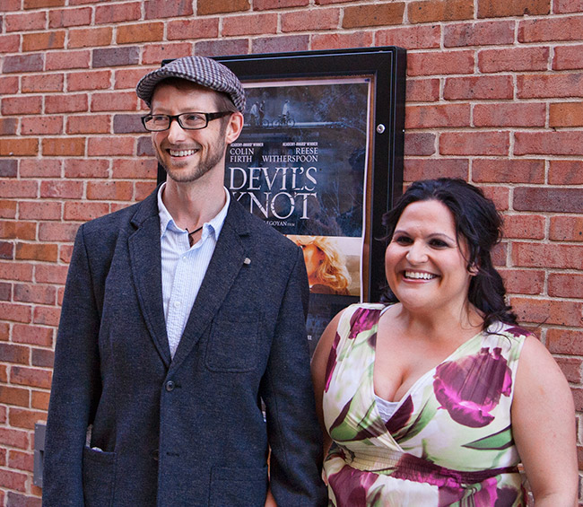White man smiling in glasses and hat standing next to white woman smiling in dress with poster on brick wall behind them