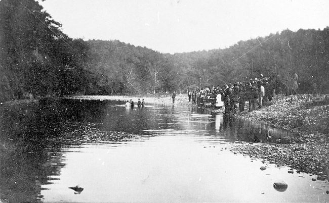 Crowd of people gathered at a tree-lined river watching a baptism