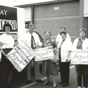 Group of white men and women with signs standing in parking lot with truck carrying a large sign behind them