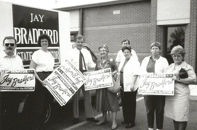 Group of white men and women with signs standing in parking lot with truck carrying a large sign behind them
