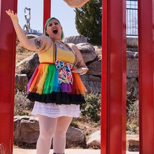White woman with tattoos and colored hair in short rainbow dress and black boots singing outdoors
