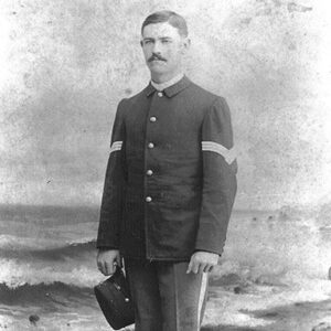 White man with side-parted dark hair and a mustache standing on beach in military uniform with cap in right hand
