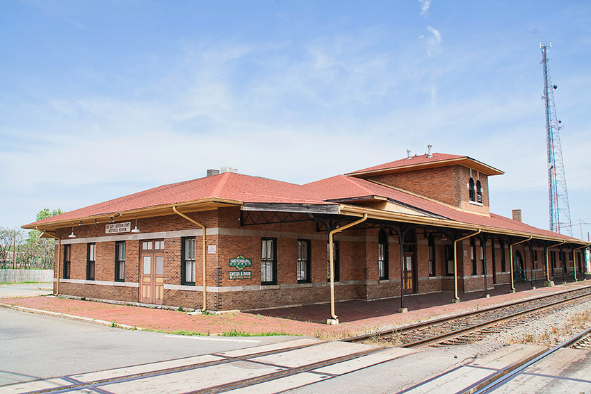 Brick depot building with covered walkway on railroad tracks with steel tower in the background