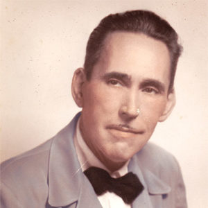 White man with pencil mustache and combed-back hair in pale suit jacket and black bow tie