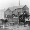 Men driving horse drawn wagons loaded with cotton outside gin facility with men standing on elevated platform in the background