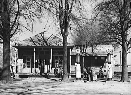Single-story gas station with pumps and three trees on dirt road
