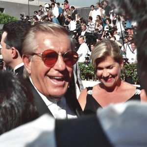 White man in suit and sunglasses and white woman in black dress at crowded outdoor event