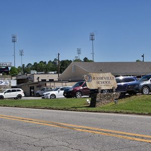 Two-lane road next to school campus with parking lot and single-story buildings with sign and stadium lights in the background