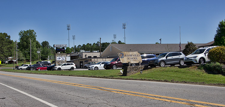 Two-lane road next to school campus with parking lot and single-story buildings with sign and stadium lights in the background