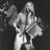 shirtless white man with long hair and tight pants holding washboard and singing into a microphone on stage