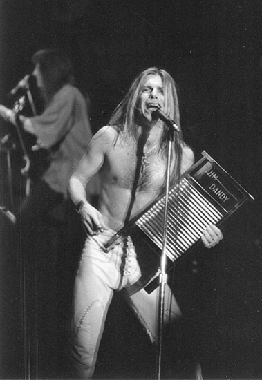 shirtless white man with long hair and tight pants holding washboard and singing into a microphone on stage