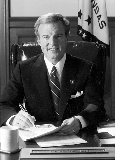White man in suit and tie smiling at his desk with Arkansas flag behind him