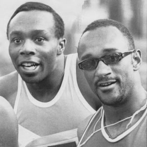 Two African-American men in Olympic track uniforms with one of the men wearing sunglasses