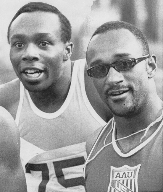 Two African-American men in Olympic track uniforms with one of the men wearing sunglasses