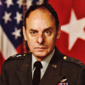 White man in military uniform with flag behind him