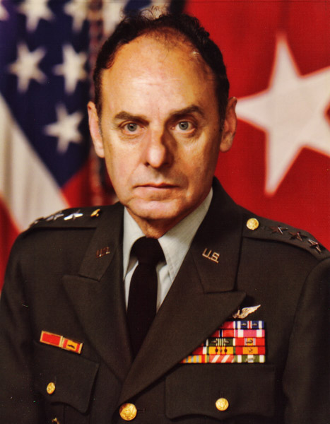 White man in military uniform with flag behind him