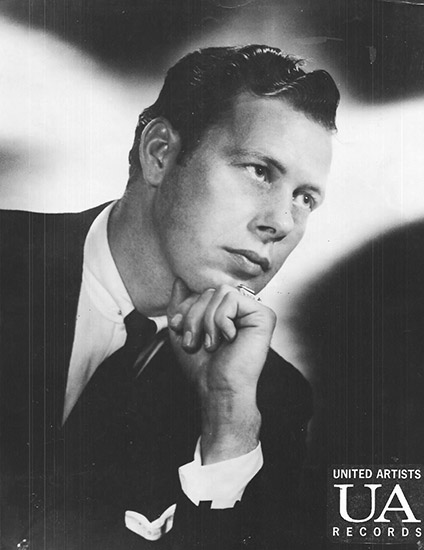 White man in suit and tie with slicked back dark hair posing with his fist under his chin