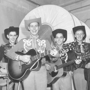 Four smiling white men in western clothing holding stringed musical instruments with fake covered wagon behind them