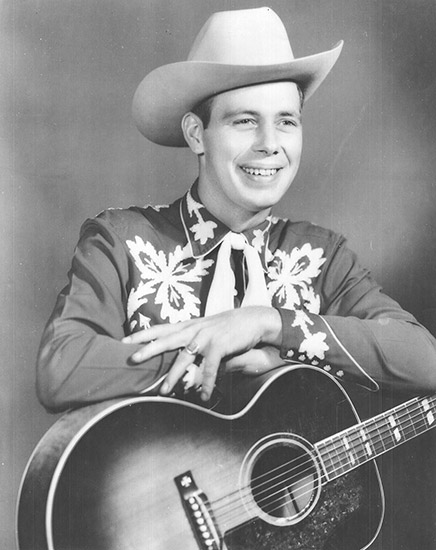 Young white man smiling in cowboy hat and western clothing posing with an acoustic guitar