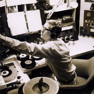 White man in long sleeved striped shirt sitting in radio booth with arm outstretched