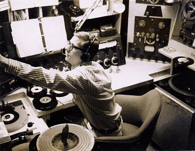 White man in long sleeved striped shirt sitting in radio booth with arm outstretched