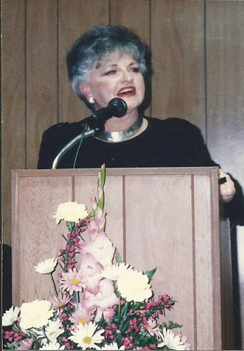 Older white woman speaking at lectern with flowers in front of it