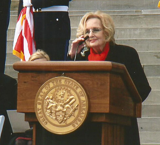 White woman with glasses and black coat speaking at lectern