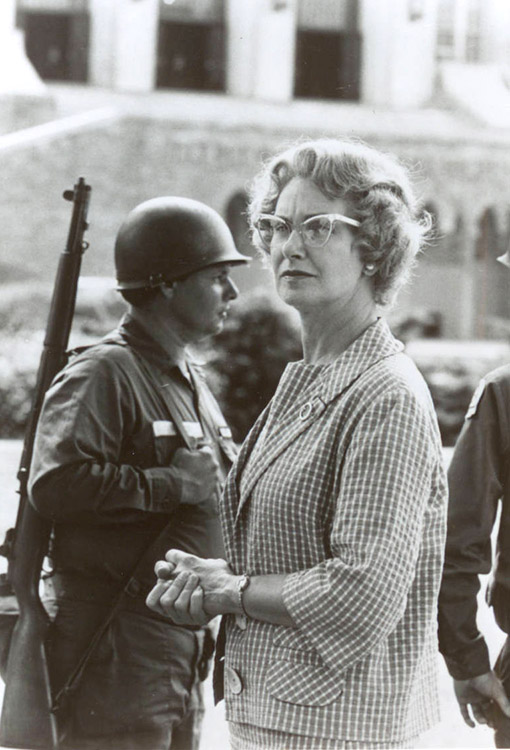 White woman with glasses in checkered shirt standing with white soldiers in uniform with guns outside school building