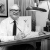 Older white man with glasses smiling at his desk with pen in his hand