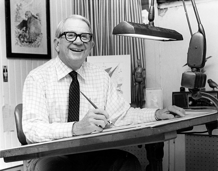 Older white man with glasses smiling at his desk with pen in his hand