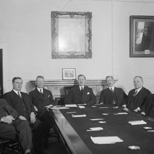 Group of seven white men in suits and ties seated at long table