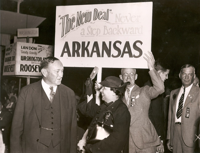 white man and woman talking in front of other white people holding political signs including "The New Deal Never a Step Backward Arkansas"