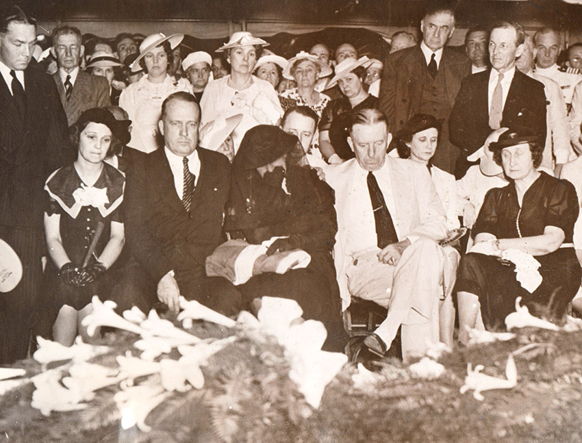 Crowd of white men and women seated and standing before flower-draped casket