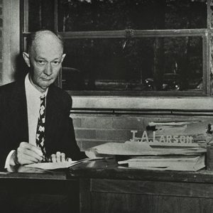 Older white man in suit and tie sitting at desk