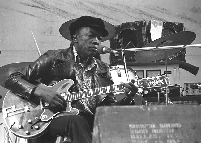 African-American man wearing black hat and playing electric guitar on stage with drums behind him and speaker in foreground