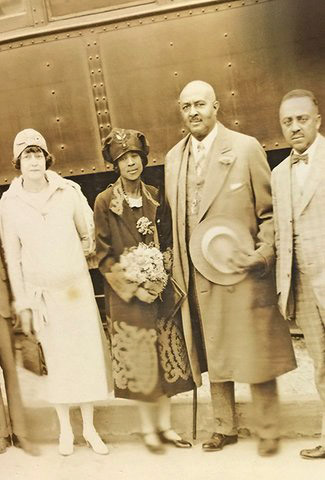 African-American man in suit holding hat standing on train platform with African-American man in suit and two women