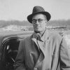 man with hat glasses scarf and coat smiling and standing by a car