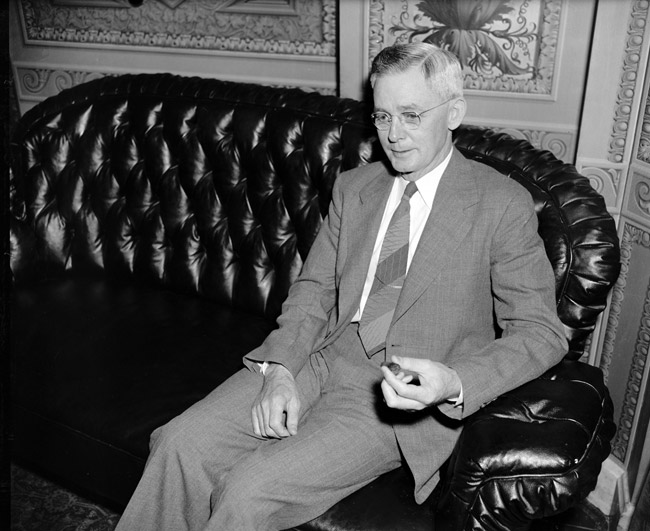 White man in glasses and suit sitting on a leather couch